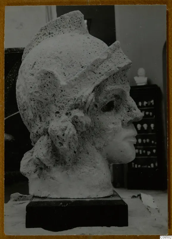 Helmeted head sculpture in pale, pitted stone, shown in right profile on a dark rectangular base