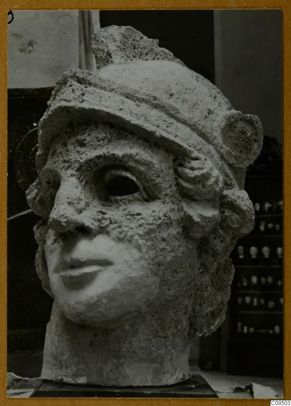 Carved stone head with a close-fitting cap, large hollow eye sockets and a damaged nose, on a dark base