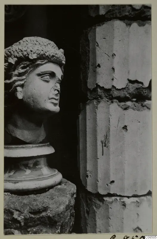 Carved stone head with wavy hair on a moulded base, set on a rough stone block beside a masonry wall