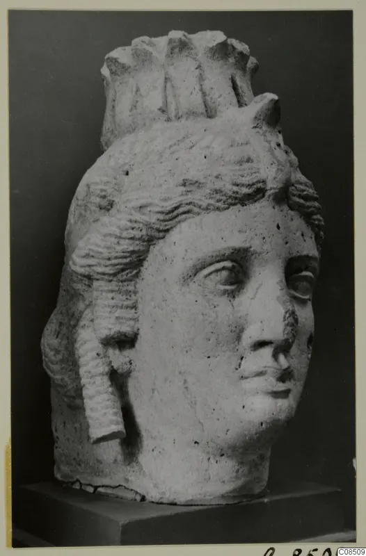 Carved stone head of a woman with wavy hair and a tall crown-like headdress, on a dark wooden stand.