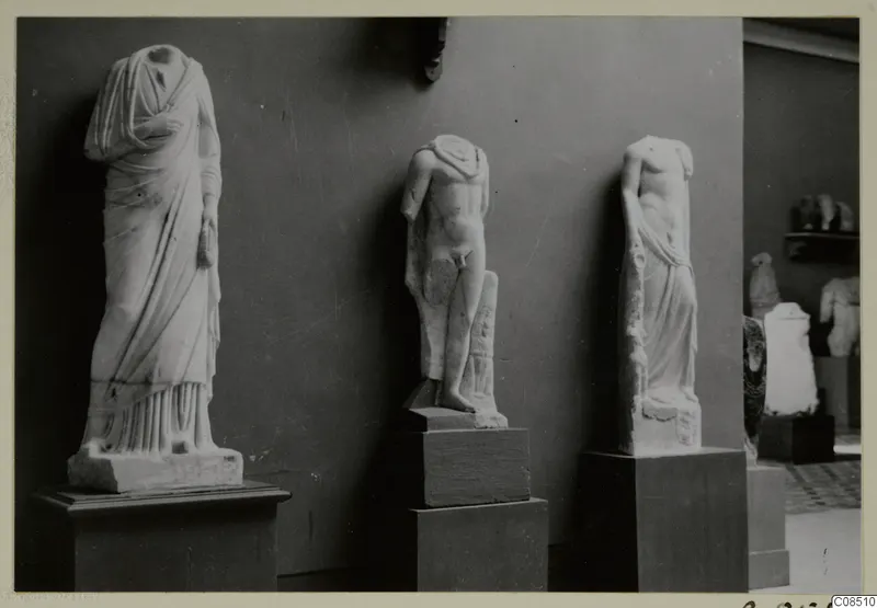 Three headless stone statues on pedestals against a dark wall, with more sculptures in a room to the right.