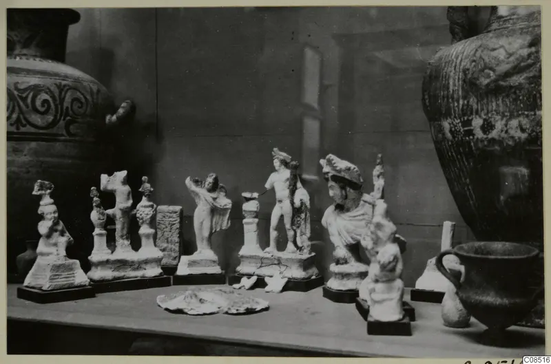 Small white figurines and a dark bowl displayed on a shelf, with large vessels behind them