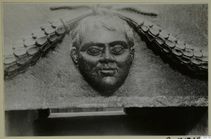 Carved stone relief of a man's face with curled hair, set between scale-patterned ornament on a block.