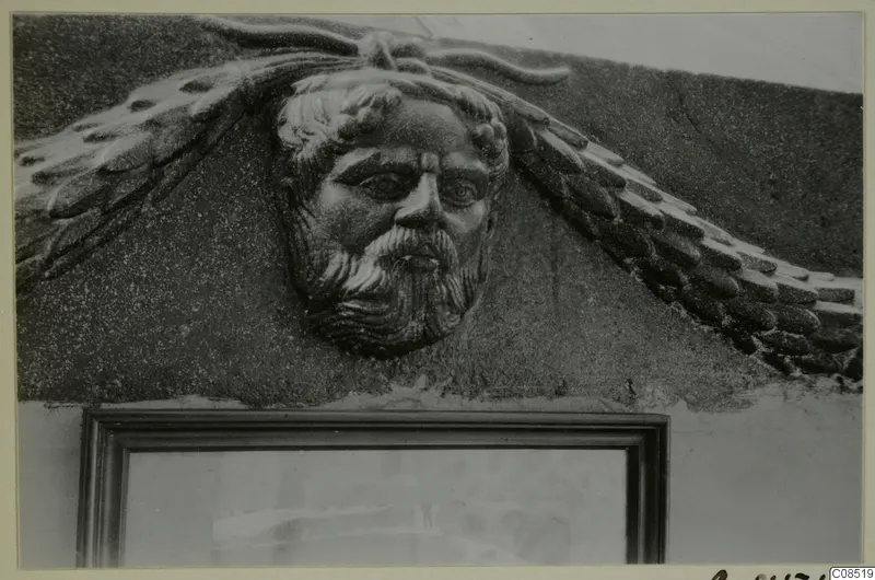 Carved bearded male face in relief, with feather-like shapes on either side, set into a wall above a framed opening