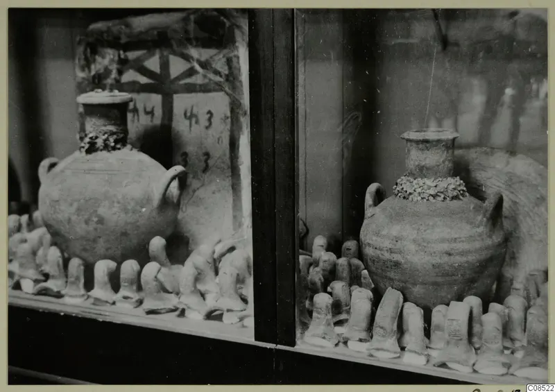 Two round jars with short necks and handles standing in a glass display case among rows of small clay objects