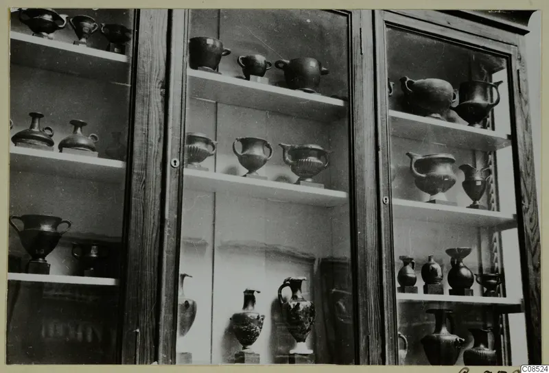 Dark vessels with handles and jugs displayed on shelves in a glass-fronted wooden cabinet.