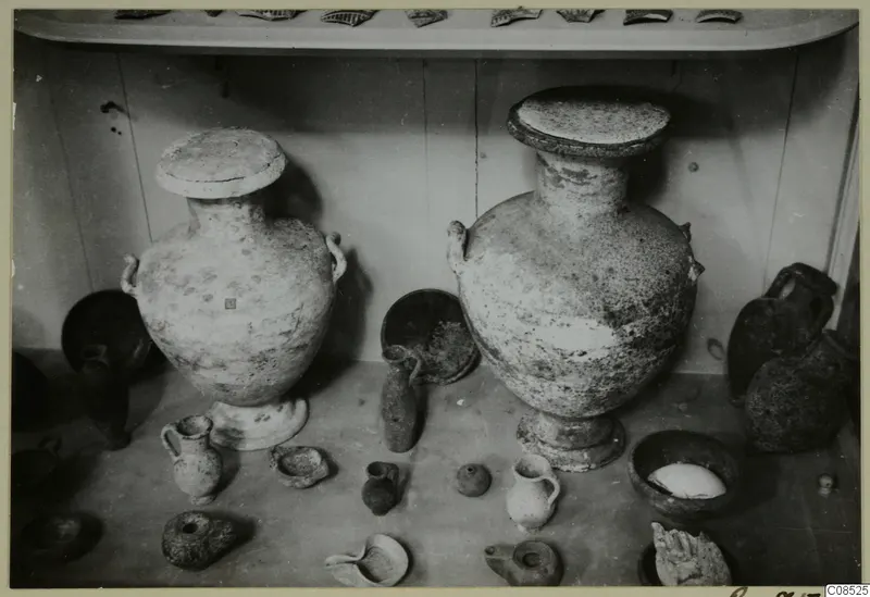 Two large two-handled jars with lids, with smaller vessels and lamps, displayed on a shelf