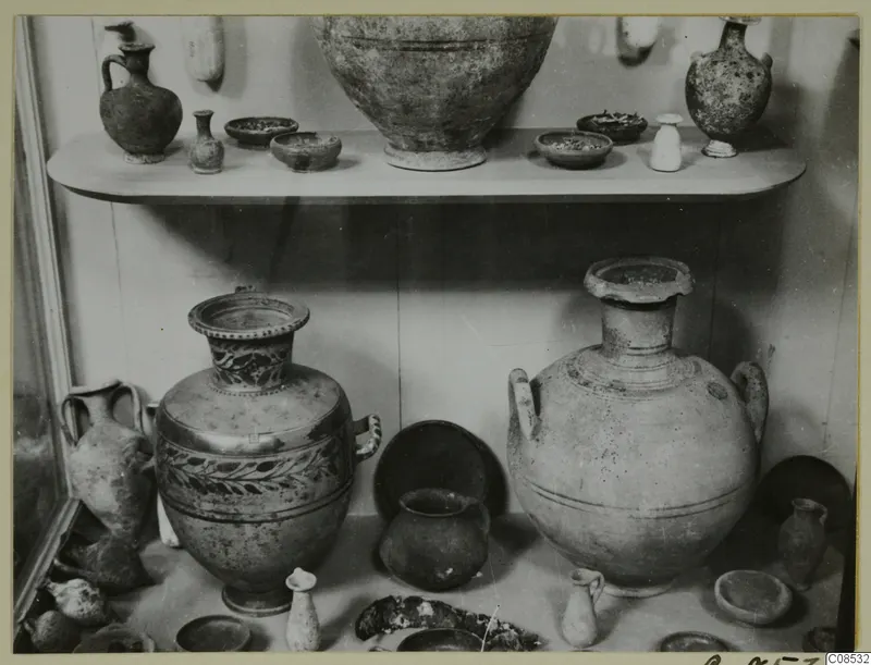 Pottery vessels displayed on two shelves in a glass case, including large two-handled jars and small juglets.