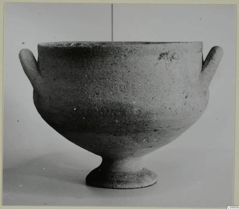 Unpainted pottery bowl with two small horizontal handles and a short footed base, photographed against a plain grey background.
