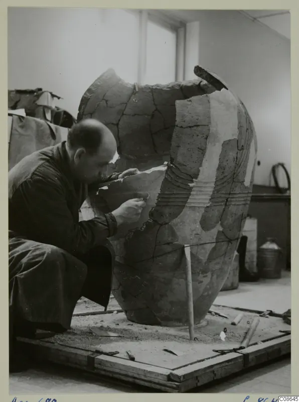 Man restoring a large broken pithos-type jar on a wooden board in a workroom