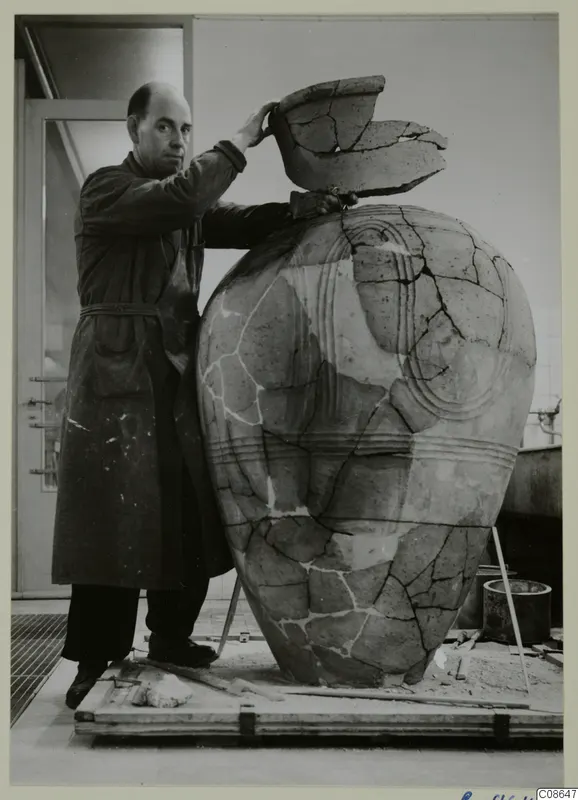Man in a dark work coat with one hand on a broken rim fragment atop a large mended pottery vessel
