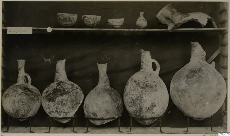 Pottery vessels displayed on a shelf: three bowls and a small juglet above, five globular juglets on wire stands below, from Lapithos.