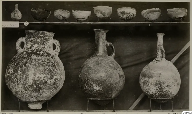 Pottery jars, a jug, a flask and a row of bowls displayed on a shelf against a dark cloth, from Lapithos Markides