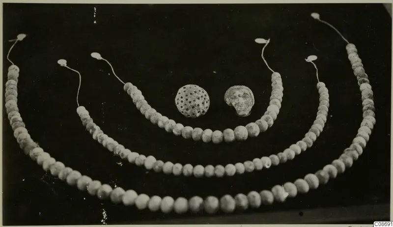 Three strands of pale beads with a perforated round pendant and a small face-shaped pendant on a dark background