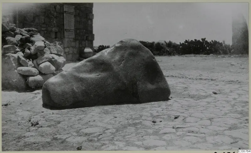 Large rounded stone block lying on a paved ground beside a stone wall, with cacti behind, at Paphos area.