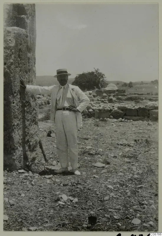Man in a light suit and boater hat standing beside a large stone block, with low stone walls behind him