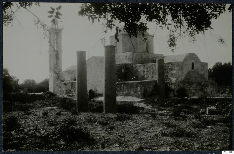 Stone building with a tall bell tower and two standing pillars in front of it, with tree branches across the top