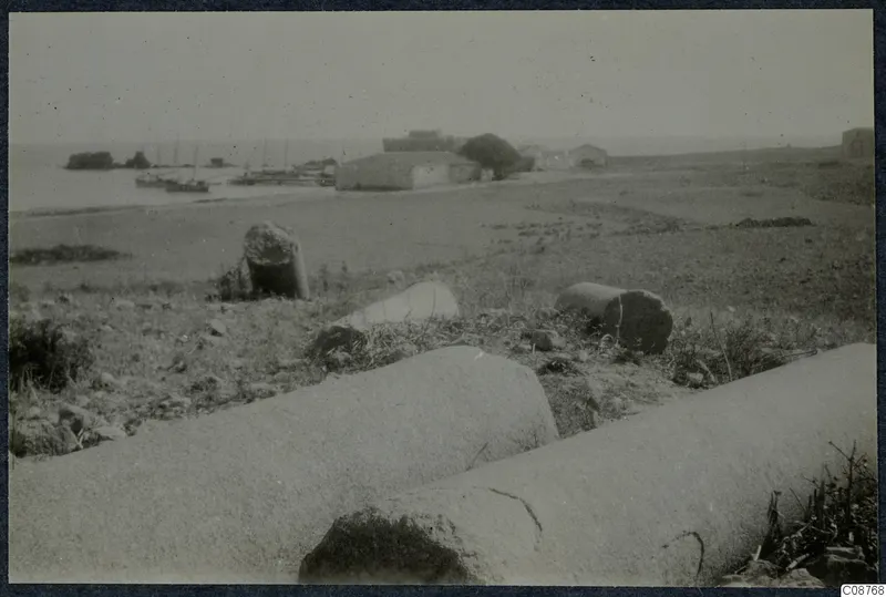 Fallen column drums in the foreground, with small boats on the water and low buildings along the shore beyond.