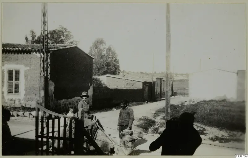Village street with a laden donkey or mule, a woman in a wide-brimmed hat and a man in a sweater beside a metal gate.