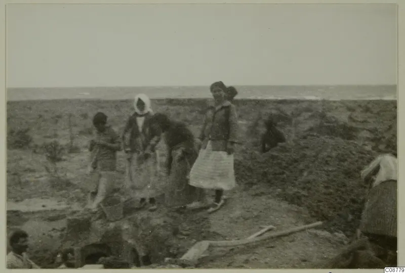 People digging beside a pit on flat ground by the sea, with a bucket and a wooden tool in the foreground