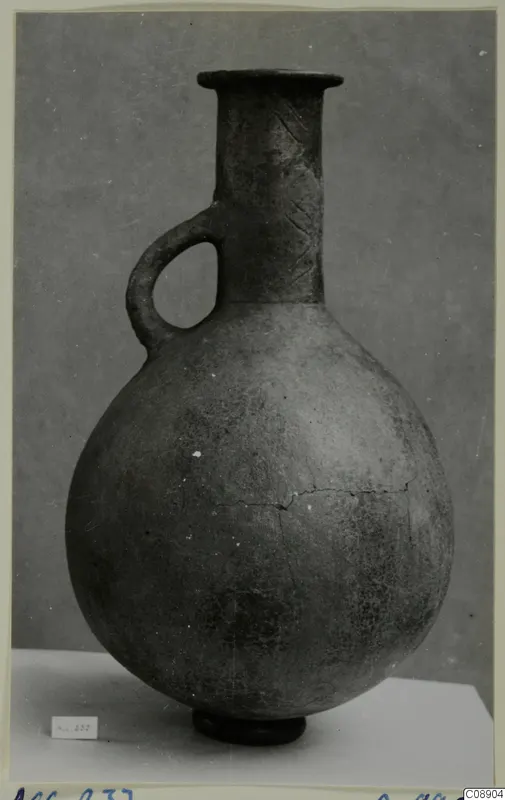 Red Polished III jug with a single loop handle and flared rim, photographed on a white plinth against a grey backdrop