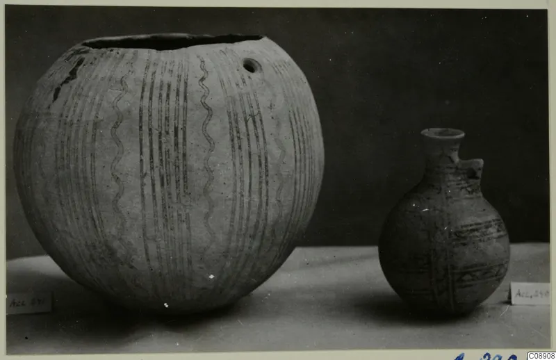 Two painted pottery vessels, a large globular jar and a small juglet, standing on a pale surface against a dark backdrop