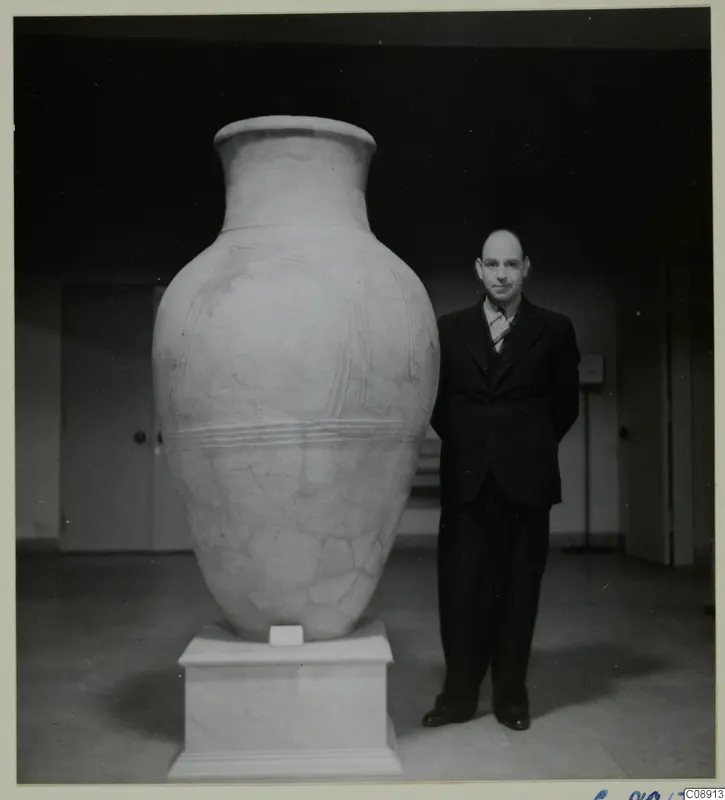Large pale pottery pithos on a white plinth, with a man in a dark suit standing beside it for scale.
