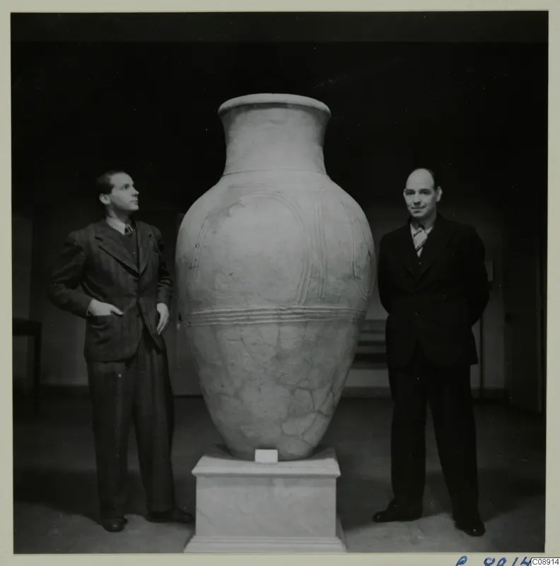 Two men stand beside a very large pottery pithos ware jar on a white pedestal in a dark room.