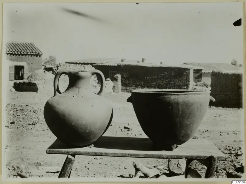 Two new clay pots on a wooden table in a village yard, with mud-brick houses behind.