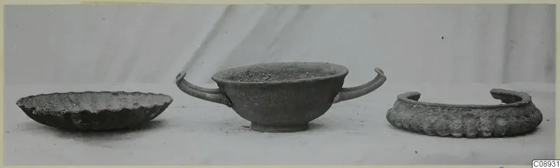Three corroded silver objects on a pale surface: a shallow scalloped dish, a two-handled cup and a wide ring-shaped object.