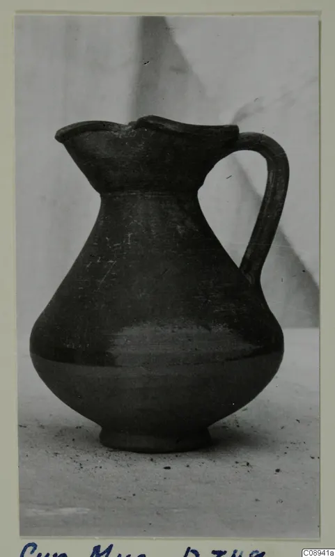 Dark jug with a pinched mouth and a single strap handle, photographed on a plain surface against a grey backdrop
