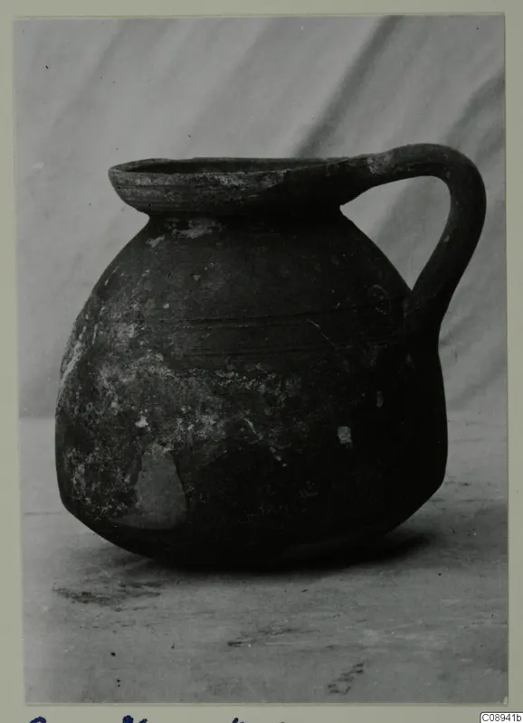 Dark handled jug with a wide flat rim and faint incised lines on the body, photographed against a grey backdrop
