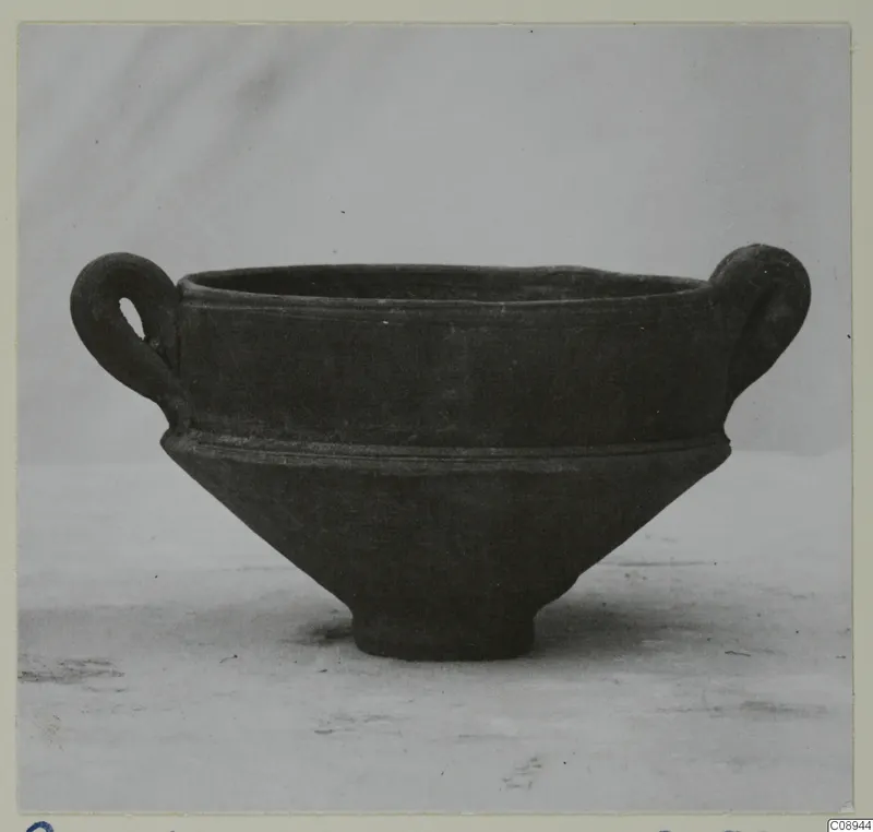 Dark grey two-handled bowl with a wide rim, a ridge at the shoulder and a short footed base, photographed on a plain surface.