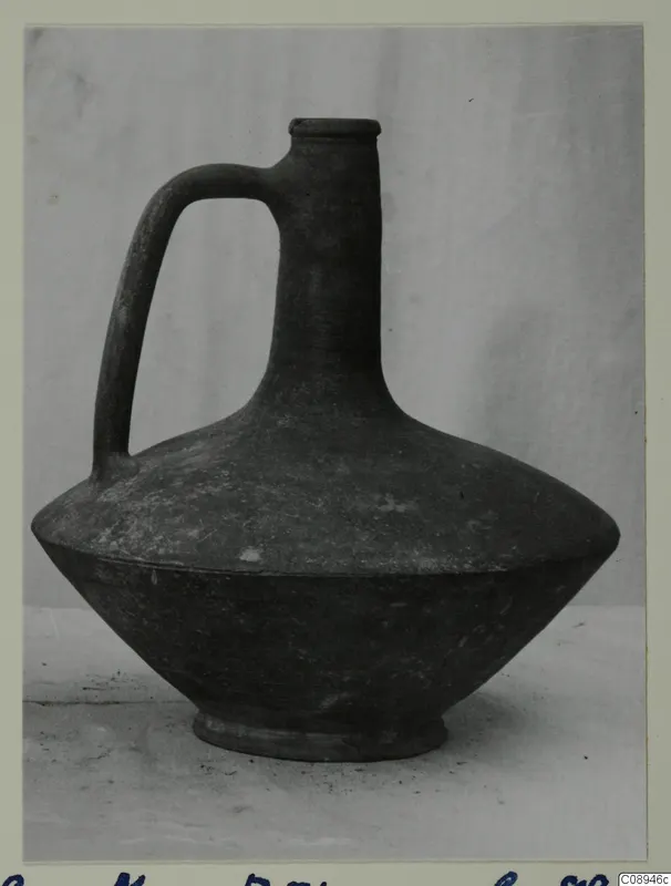 Single-handled pottery jug with a narrow neck, flat rim and wide, sloping body, shown on a plain backdrop