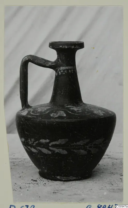 Dark-surfaced juglet with a single strap handle and pale painted decoration, photographed on a mounted card