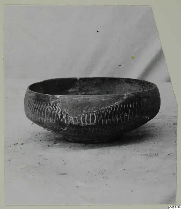 Dark bowl with bands of pale incised strokes on its body, shown on a plain surface against a light backdrop