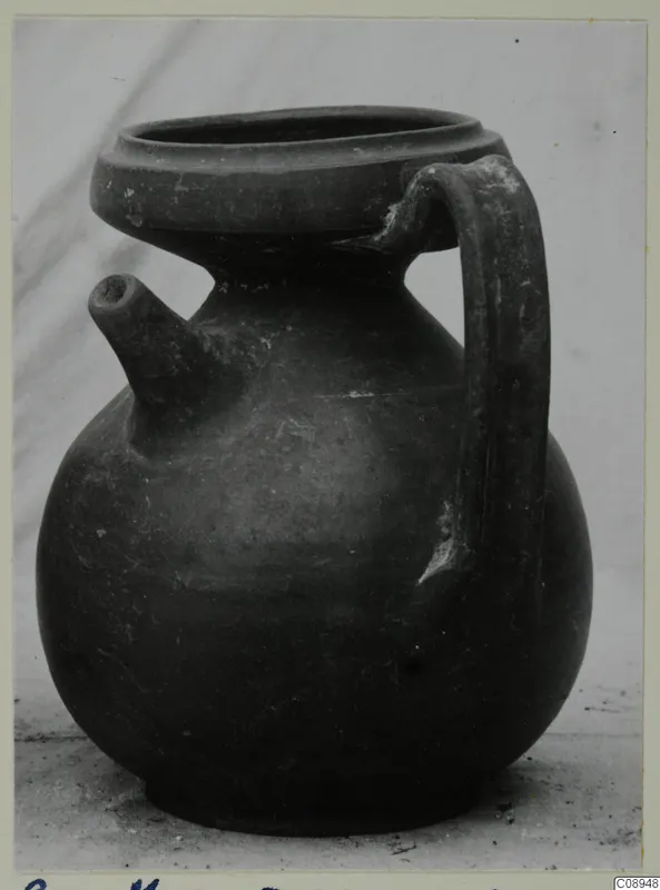 Dark globular jug with a side spout and a strap handle, shown on a pale surface