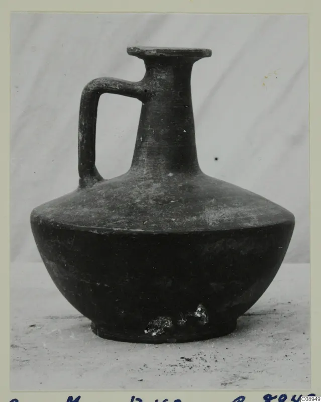 One-handled dark jug with a flared rim, narrow neck and ring base, shown against a grey backdrop.