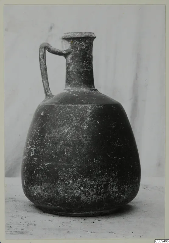 Dark jug with a single loop handle, narrow neck and flaring rim, photographed against a pale backdrop