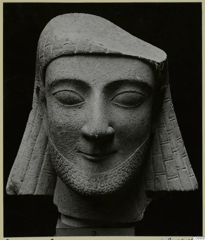 Limestone head of a bearded figure in a close-fitting headdress with side flaps, set on a numbered base