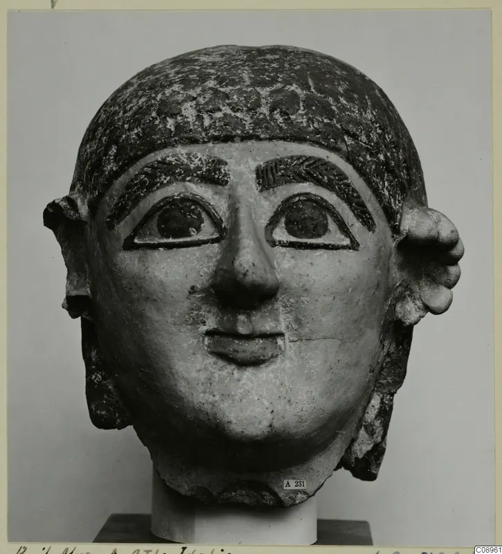 Terracotta head with a painted hairstyle, large ears and a calm face, shown on a stand.