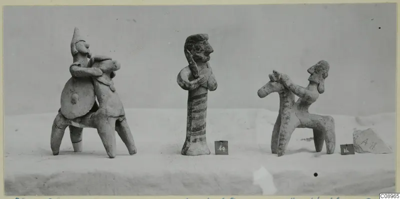 Three terracotta figurines on a pale cloth: two riders on four-legged forms and a standing figure with a striped body.