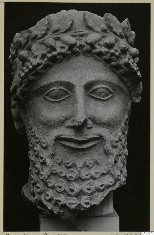 Limestone head of a bearded male figure with a wreath of leaves and berries, shown against a dark background.