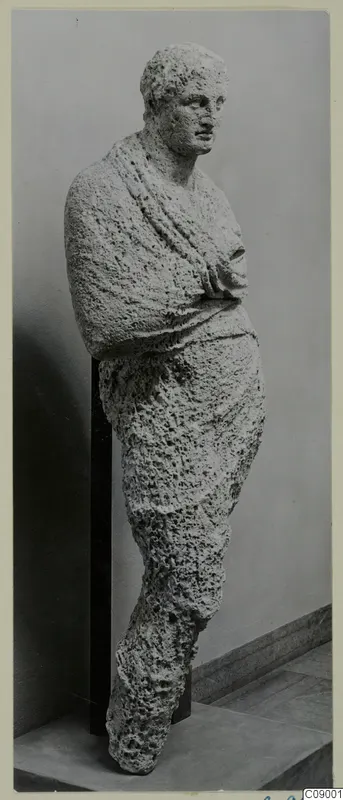 Full-length statue of a standing figure in rough, pitted limestone, with a draped cloak and arms folded at the chest
