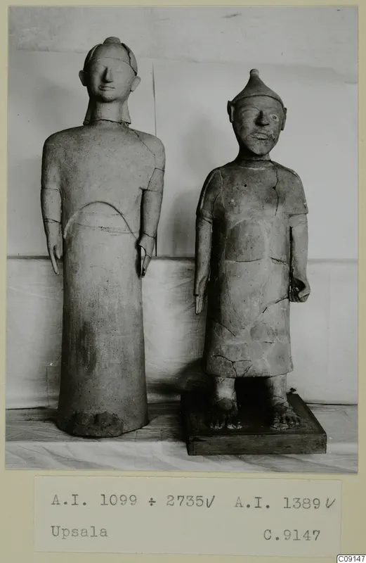 Two standing clay statues of human figures, one on a dark wooden base, against a pale draped backdrop