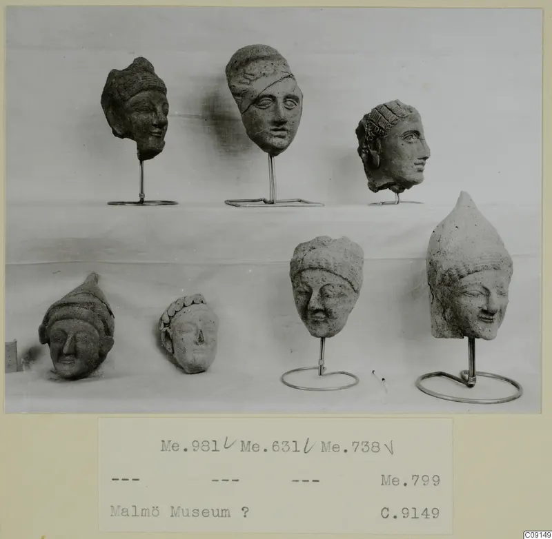 Seven small clay heads of human figures on a cloth-covered shelf, five on thin wire stands, in a photograph mounted on a card