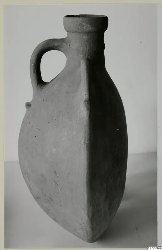 Pottery flask with a loop handle and a flattened body, standing on a pale surface against a grey backdrop.