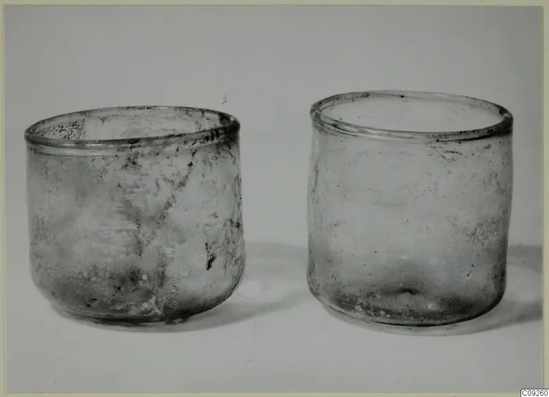 Two blown glass beakers with weathered, clouded surfaces standing side by side on a pale background.
