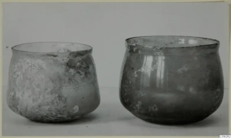 Two blown glass vessels standing side by side on a pale surface, one wider with a flared rim, one rounder and deeper.