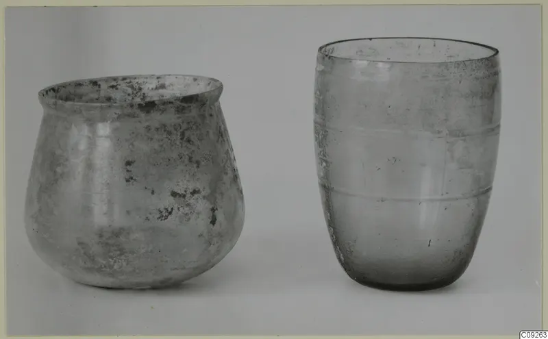 Two blown glass vessels side by side on a pale background: a squat, weathered jar left and a tall beaker with ridges right.
